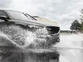 Un'auto scura attraversa una pozzanghera, sollevando un'enorme onda d'acqua. Il fenomeno dell'aquaplaning aumenta con la pioggia.