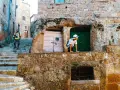 Un ragazzo con cappello bianco sporge da un muro di tufo in una stretta via di una città del tufo toscana. Un uomo con zaino sale le scale.