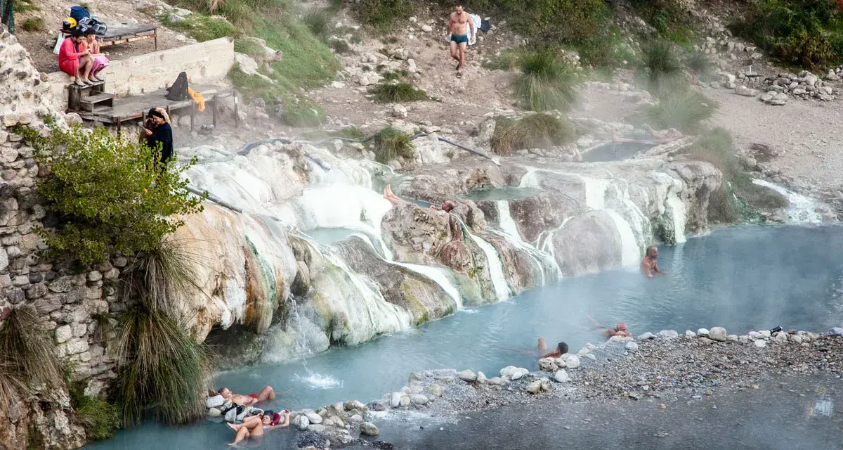 Persone si rilassano nelle calde acque delle terme di Petriolo libere, tra rocce e vegetazione.