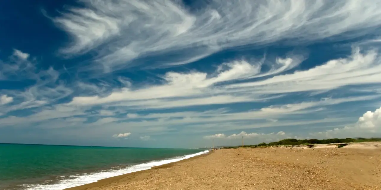 Spiaggia dorata e mare turchese al parco costiero di Rimigliano, con un cielo blu intenso solcato da nuvole bianche e leggere.