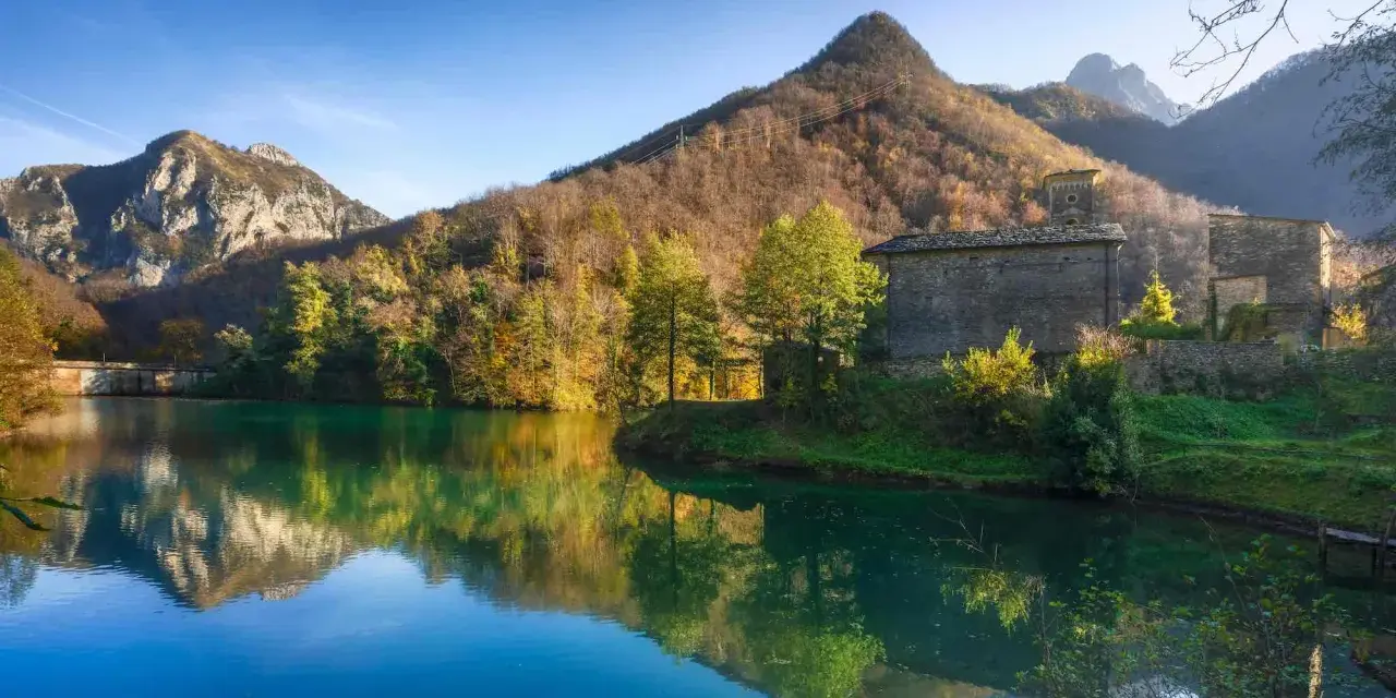 Lago incantevole con riflessi di montagne e boschi autunnali. Un borgo in pietra completa il paesaggio, ideale per scoprire dove andare in montagna in Toscana.