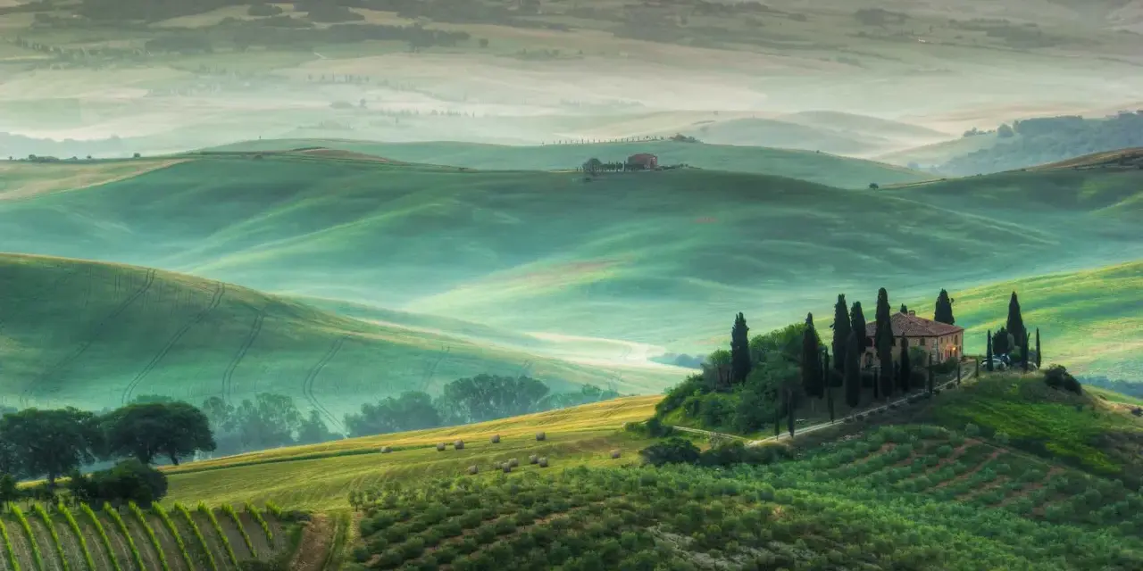 Dolci colline verdi e nebbiolina mattutina creano un'atmosfera magica nei punti panoramici della Val d'Orcia. Una casa colonica con cipressi domina la scena.