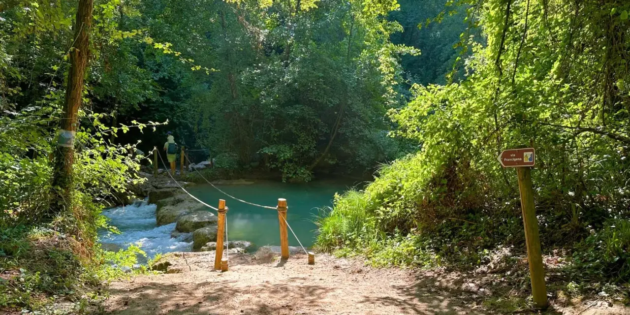 Sentiero naturalistico con guado su fiume, ideale per percorsi trekking Toscana. Segnale Via Francigena indica la direzione.