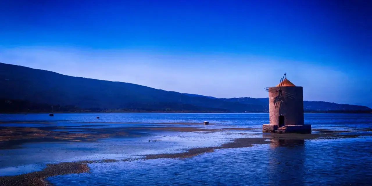 Mulino solitario in un'oasi WWF Toscana, con acque blu e colline sullo sfondo.