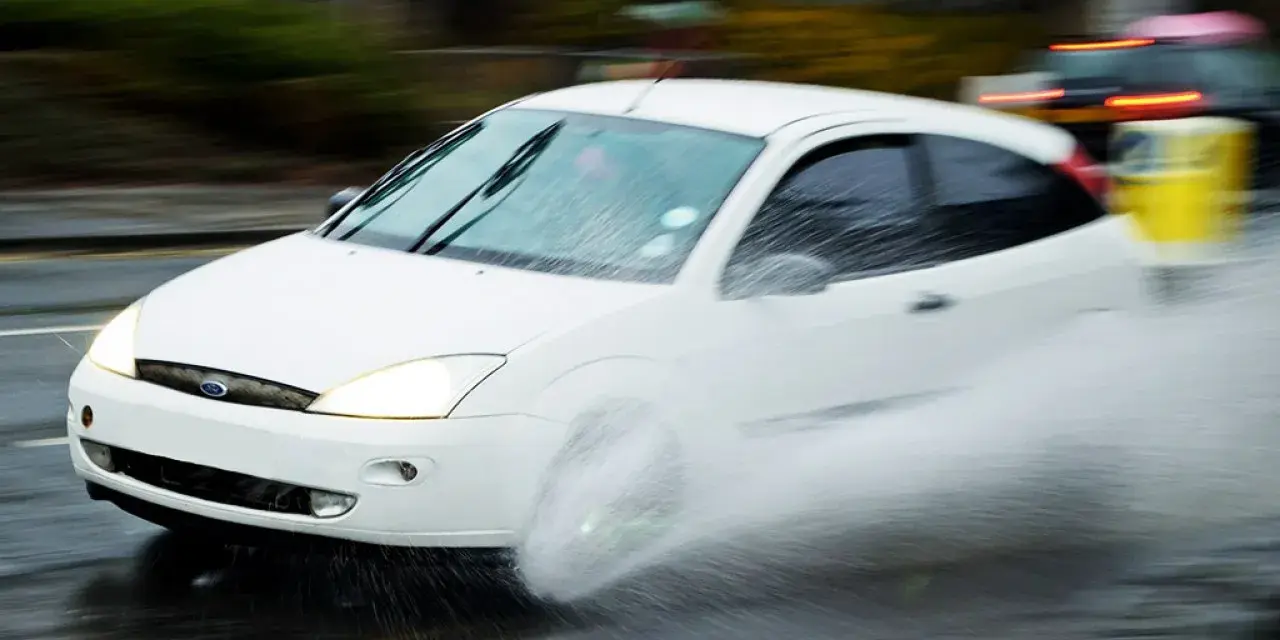 Auto bianca in movimento su strada bagnata, creando un'onda d'acqua a causa del fenomeno aquaplaning.