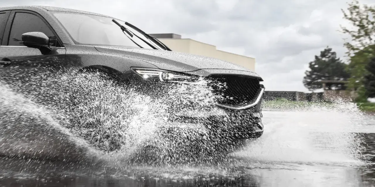 Un'auto scura attraversa una pozzanghera, sollevando un'enorme onda d'acqua. Il fenomeno dell'aquaplaning aumenta con la pioggia.