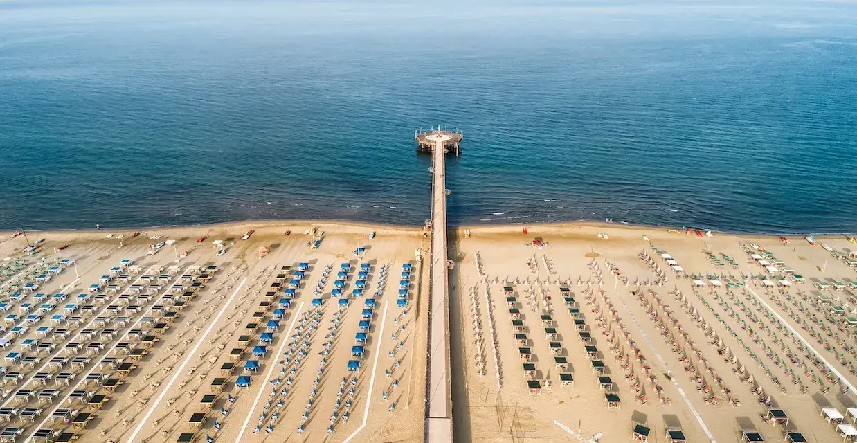 Un lungo molo si estende verso il mare calmo, con file ordinate di ombrelloni e lettini che attendono i bagnanti. Un'oasi di relax, quasi un mare vicino Firenze, dove la sabbia dorata incontra l'azzurro infinito.