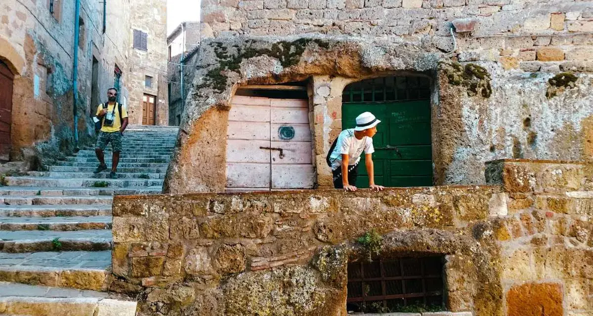Un ragazzo con cappello bianco sporge da un muro di tufo in una stretta via di una città del tufo toscana. Un uomo con zaino sale le scale.