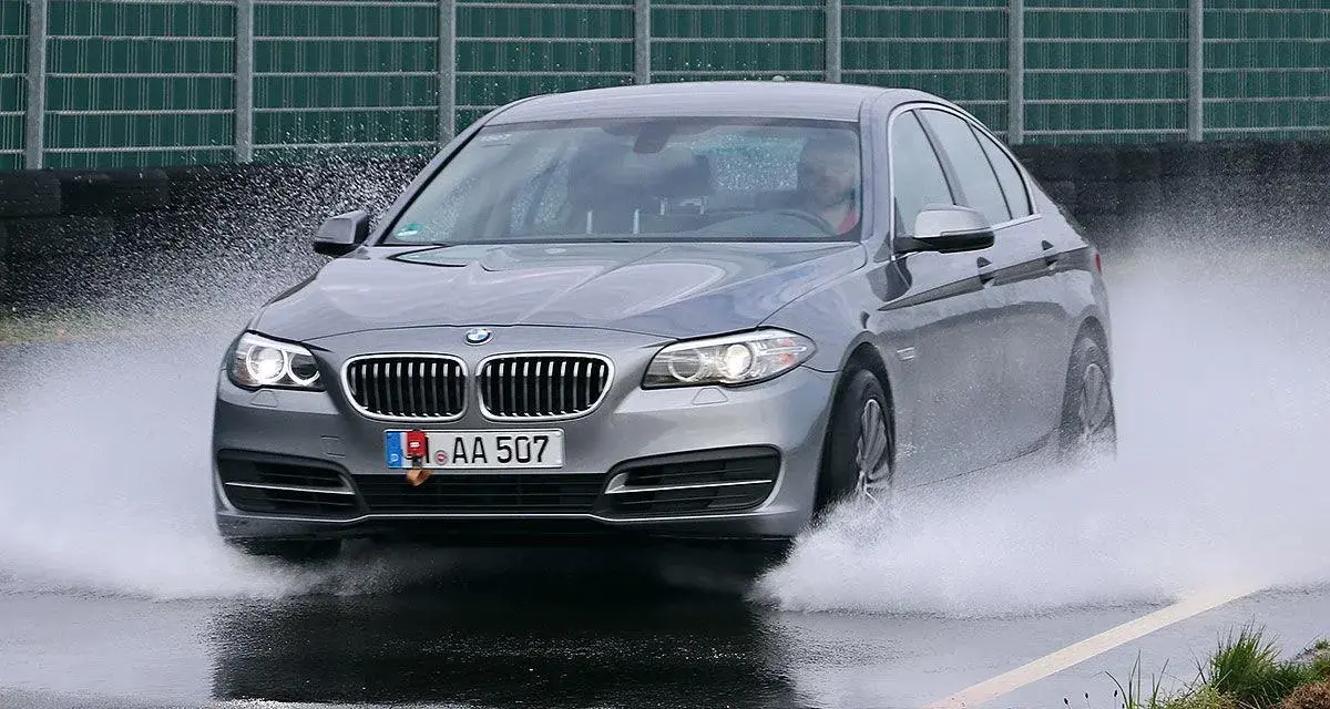 BMW grigia in test di acquaplaning, solleva un'onda d'acqua mentre sfreccia sull'asfalto bagnato.