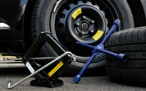 Attrezzi per cambiare una ruota: cric, chiave a stella blu e ruotino di scorta vicino a un veicolo.