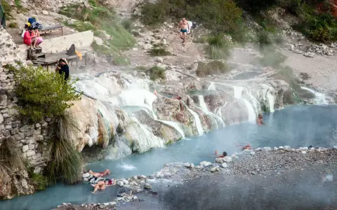 Persone si rilassano nelle calde acque delle terme di Petriolo libere, tra rocce e vegetazione.