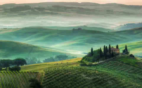 Dolci colline verdi e nebbiolina mattutina creano un'atmosfera magica nei punti panoramici della Val d'Orcia. Una casa colonica con cipressi domina la scena.