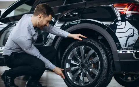 Uomo in ginocchio effettua il controllo gomme auto, esaminando il battistrada del pneumatico nero.