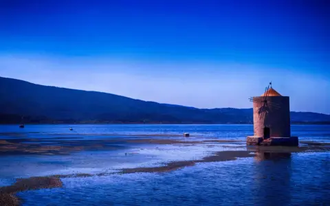 Mulino solitario in un'oasi WWF Toscana, con acque blu e colline sullo sfondo.