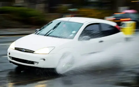 Auto bianca in movimento su strada bagnata, creando un'onda d'acqua a causa del fenomeno aquaplaning.