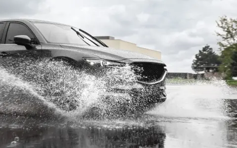 Un'auto scura attraversa una pozzanghera, sollevando un'enorme onda d'acqua. Il fenomeno dell'aquaplaning aumenta con la pioggia.