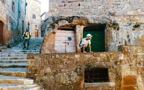 Un ragazzo con cappello bianco sporge da un muro di tufo in una stretta via di una città del tufo toscana. Un uomo con zaino sale le scale.