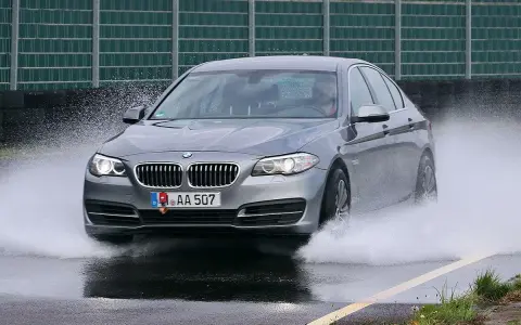 BMW grigia in test di acquaplaning, solleva un'onda d'acqua mentre sfreccia sull'asfalto bagnato.