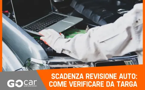 Meccanico con guanti controlla la scadenza della revisione auto tramite laptop, con il cofano aperto.