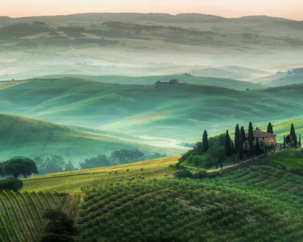 Dolci colline verdi e nebbiolina mattutina creano un'atmosfera magica nei punti panoramici della Val d'Orcia. Una casa colonica con cipressi domina la scena.