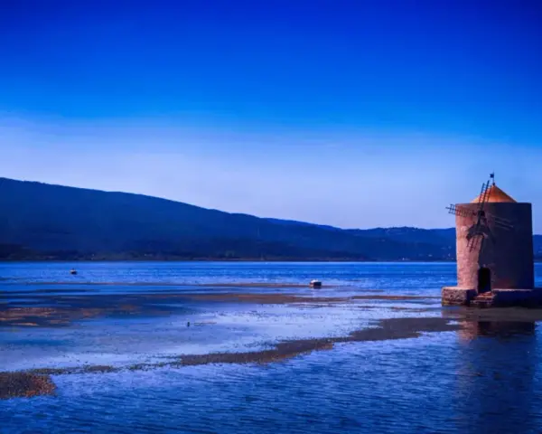 Mulino solitario in un'oasi WWF Toscana, con acque blu e colline sullo sfondo.