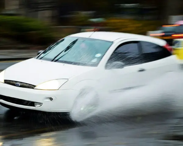 Auto bianca in movimento su strada bagnata, creando un'onda d'acqua a causa del fenomeno aquaplaning.