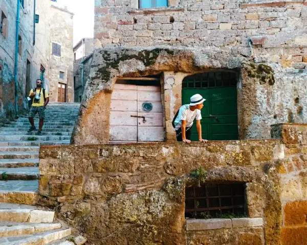 Un ragazzo con cappello bianco sporge da un muro di tufo in una stretta via di una città del tufo toscana. Un uomo con zaino sale le scale.