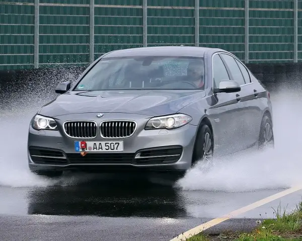 BMW grigia in test di acquaplaning, solleva un'onda d'acqua mentre sfreccia sull'asfalto bagnato.