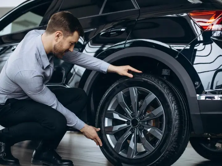 Uomo in ginocchio effettua il controllo gomme auto, esaminando il battistrada del pneumatico nero.