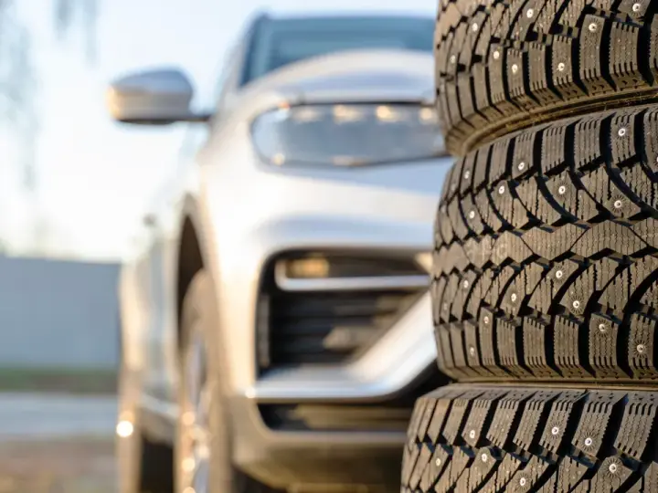 Gomme invernali con chiodi in primo piano, un'auto bianca sfocata sullo sfondo. Preparazione per l'inverno, anche se è estate.