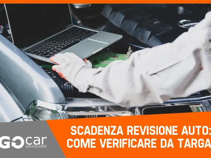 Meccanico con guanti controlla la scadenza della revisione auto tramite laptop, con il cofano aperto.