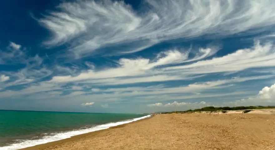 Spiaggia dorata e mare turchese al parco costiero di Rimigliano, con un cielo blu intenso solcato da nuvole bianche e leggere.