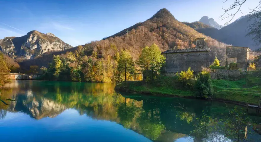 Lago incantevole con riflessi di montagne e boschi autunnali. Un borgo in pietra completa il paesaggio, ideale per scoprire dove andare in montagna in Toscana.