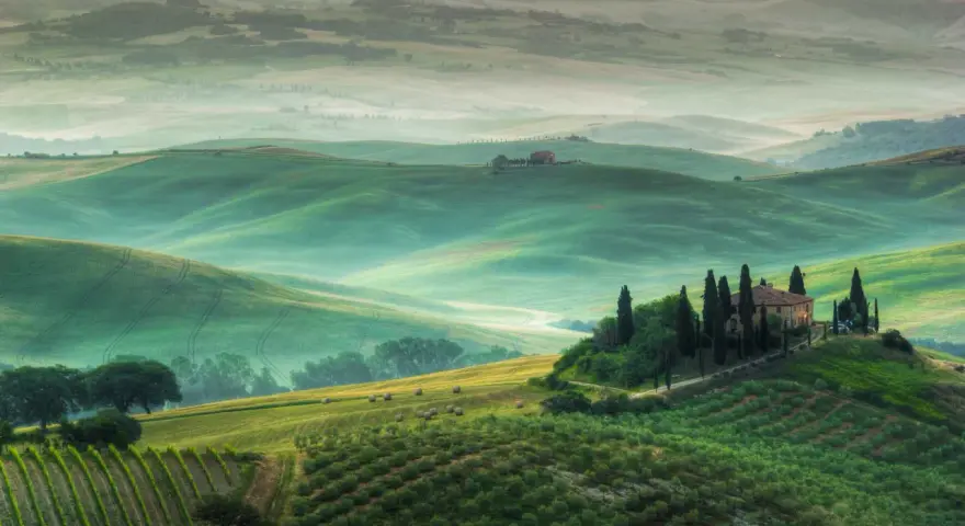 Dolci colline verdi e nebbiolina mattutina creano un'atmosfera magica nei punti panoramici della Val d'Orcia. Una casa colonica con cipressi domina la scena.