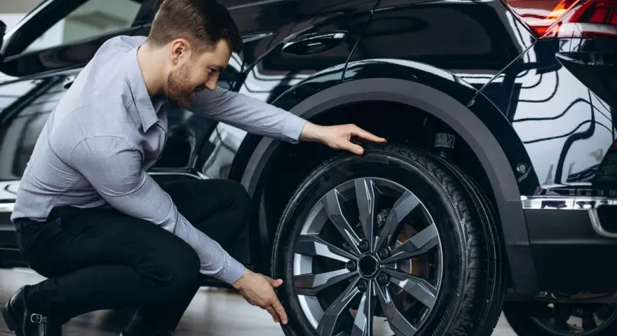 Uomo in ginocchio effettua il controllo gomme auto, esaminando il battistrada del pneumatico nero.