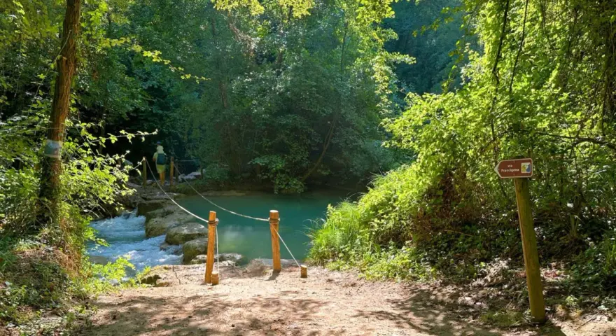 Sentiero naturalistico con guado su fiume, ideale per percorsi trekking Toscana. Segnale Via Francigena indica la direzione.