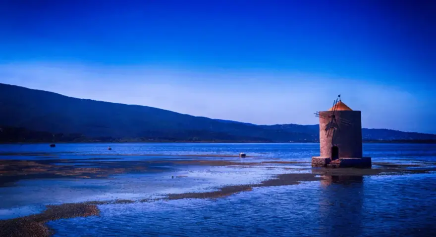 Mulino solitario in un'oasi WWF Toscana, con acque blu e colline sullo sfondo.