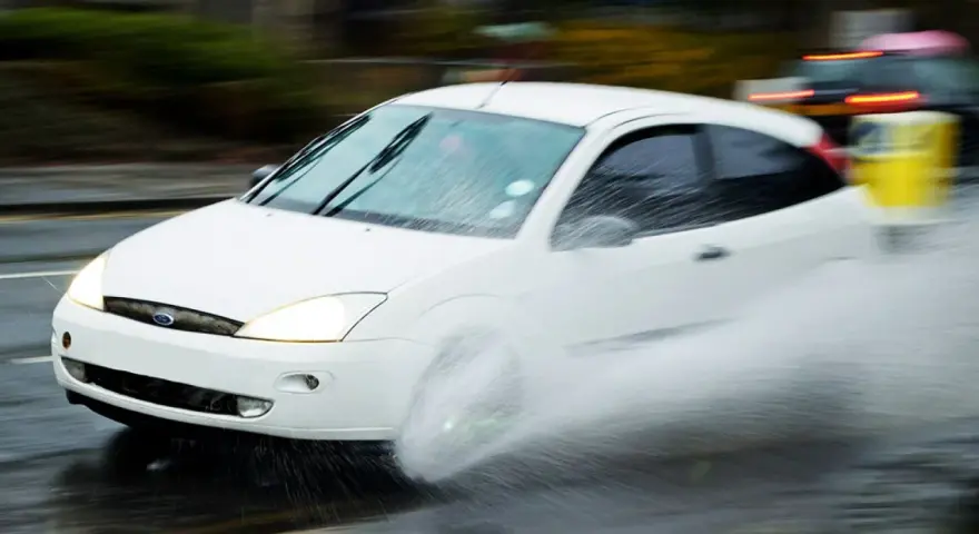 Auto bianca in movimento su strada bagnata, creando un'onda d'acqua a causa del fenomeno aquaplaning.