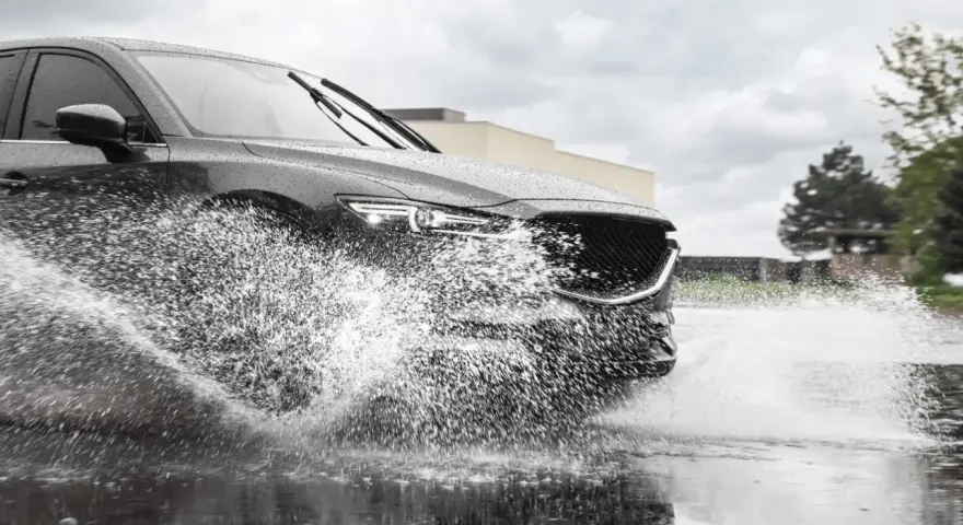 Un'auto scura attraversa una pozzanghera, sollevando un'enorme onda d'acqua. Il fenomeno dell'aquaplaning aumenta con la pioggia.
