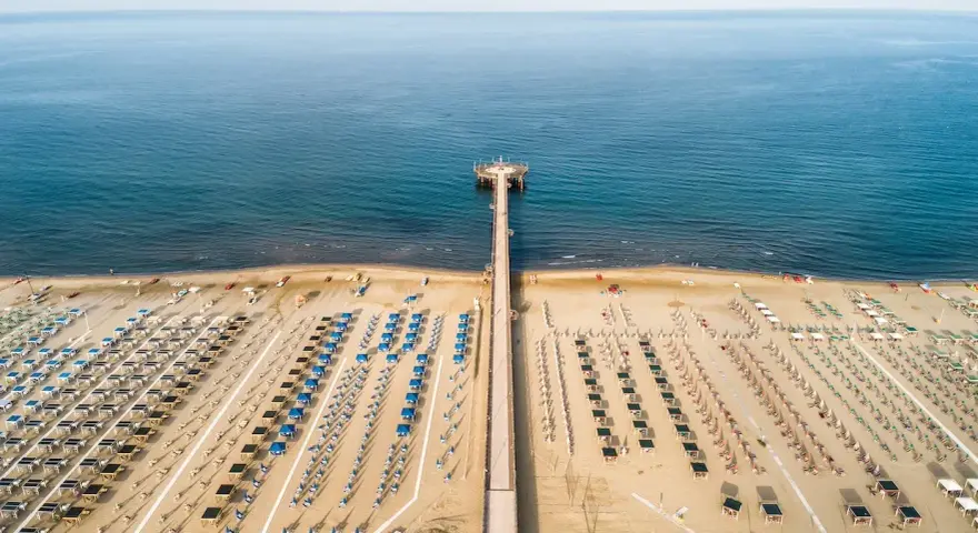 Un lungo molo si estende verso il mare calmo, con file ordinate di ombrelloni e lettini che attendono i bagnanti. Un'oasi di relax, quasi un mare vicino Firenze, dove la sabbia dorata incontra l'azzurro infinito.