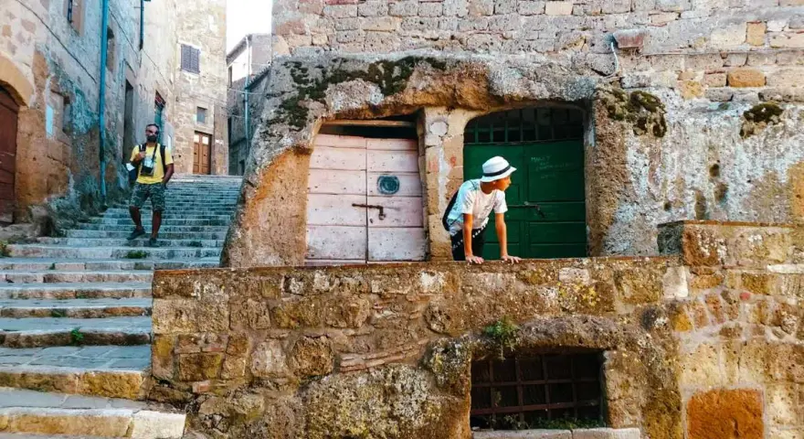 Un ragazzo con cappello bianco sporge da un muro di tufo in una stretta via di una città del tufo toscana. Un uomo con zaino sale le scale.