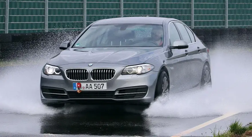 BMW grigia in test di acquaplaning, solleva un'onda d'acqua mentre sfreccia sull'asfalto bagnato.