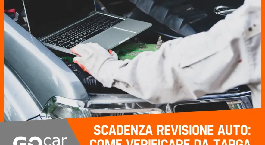 Meccanico con guanti controlla la scadenza della revisione auto tramite laptop, con il cofano aperto.