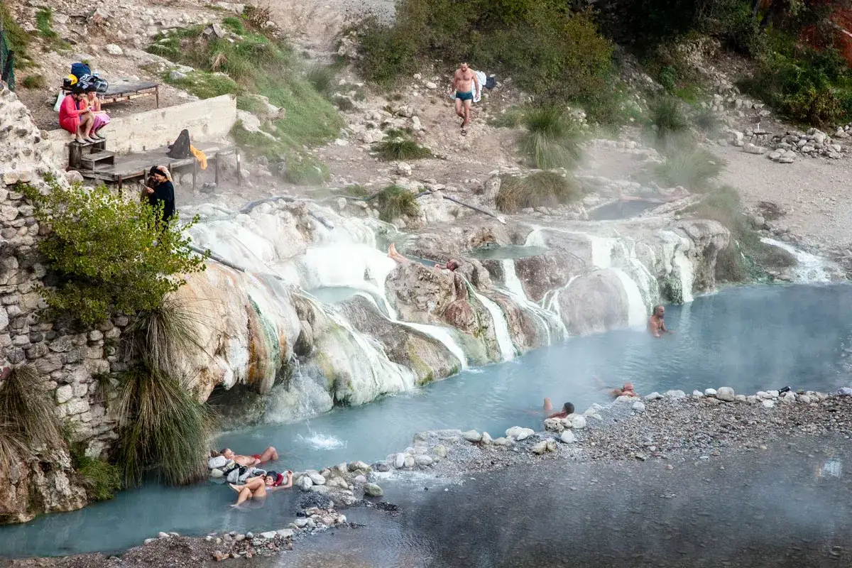 Persone si rilassano nelle calde acque delle terme di Petriolo libere, tra rocce e cascate naturali.