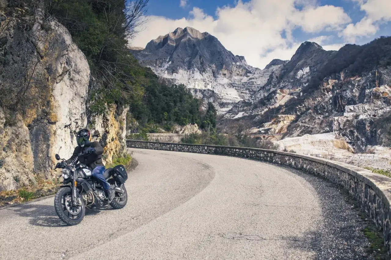 Un motociclista percorre una strada tortuosa tra le montagne della Toscana, con cave di marmo sullo sfondo.