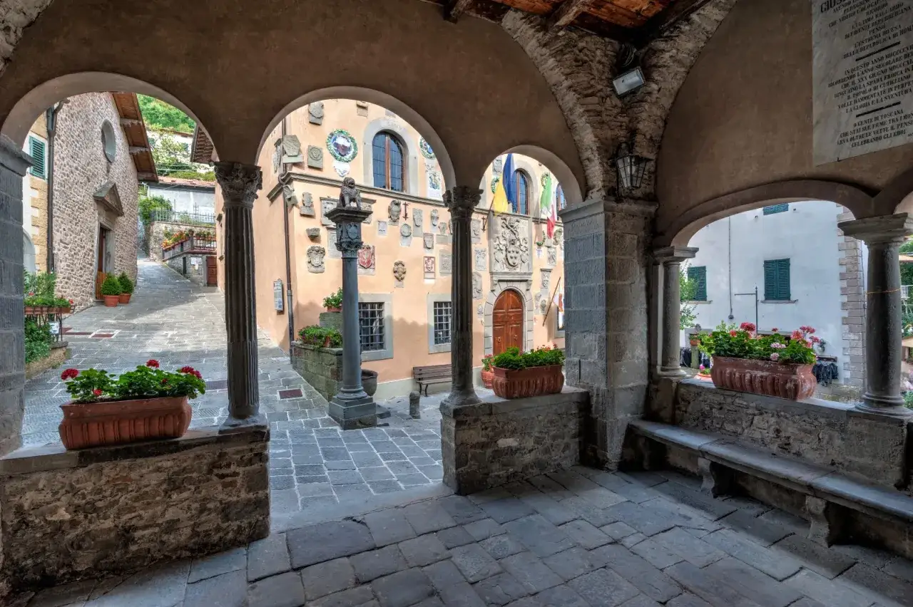Un vicolo tra i borghi della montagna pistoiese, con archi in pietra, fiori rossi e stemmi antichi su un palazzo.
