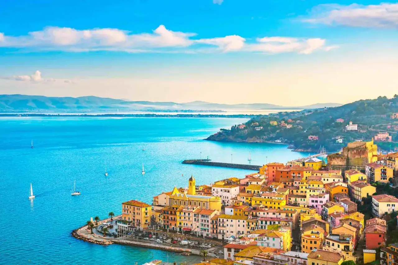Villaggio colorato affacciato sul mare che bagna la Toscana, con barche a vela che solcano le acque azzurre sotto un cielo limpido.