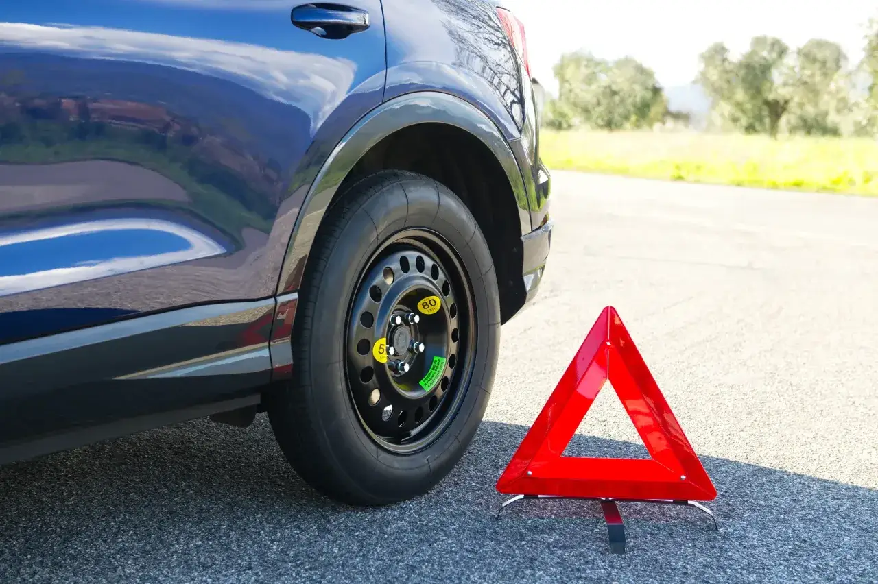 Un'auto blu ha una ruota di scorta, un ruotino, montata. Accanto, un triangolo rosso di emergenza segnala un guasto.
