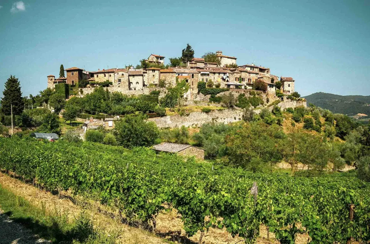 Borgo medievale su una collina, circondato da vigneti e ulivi. Le bandiere arancioni Toscana sventolano tra le case di pietra.