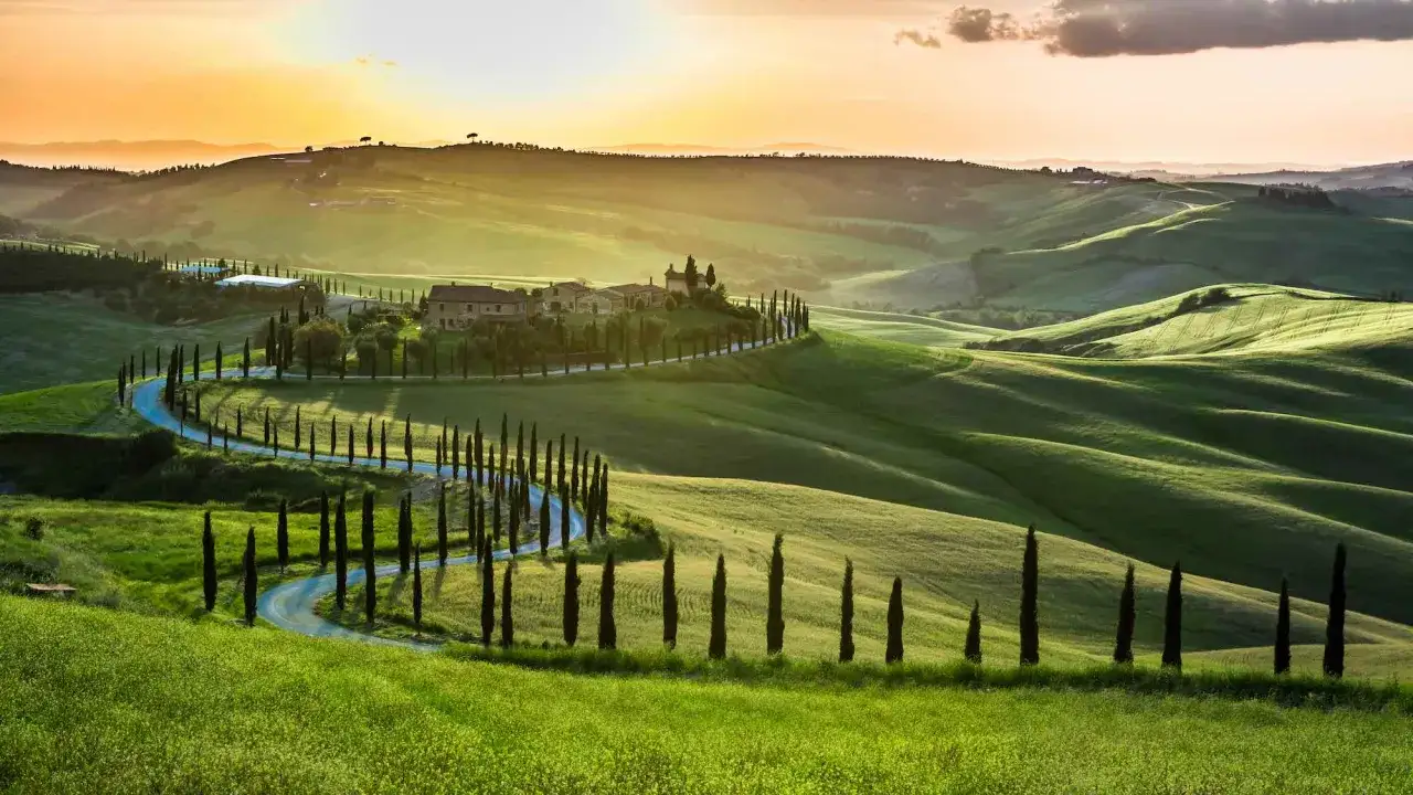 Dolci colline verdi toscane con cipressi lungo una strada tortuosa, un casale in cima e il sole al tramonto. Un paesaggio da cartolina delle citt&agrave; pi&ugrave; belle toscana.